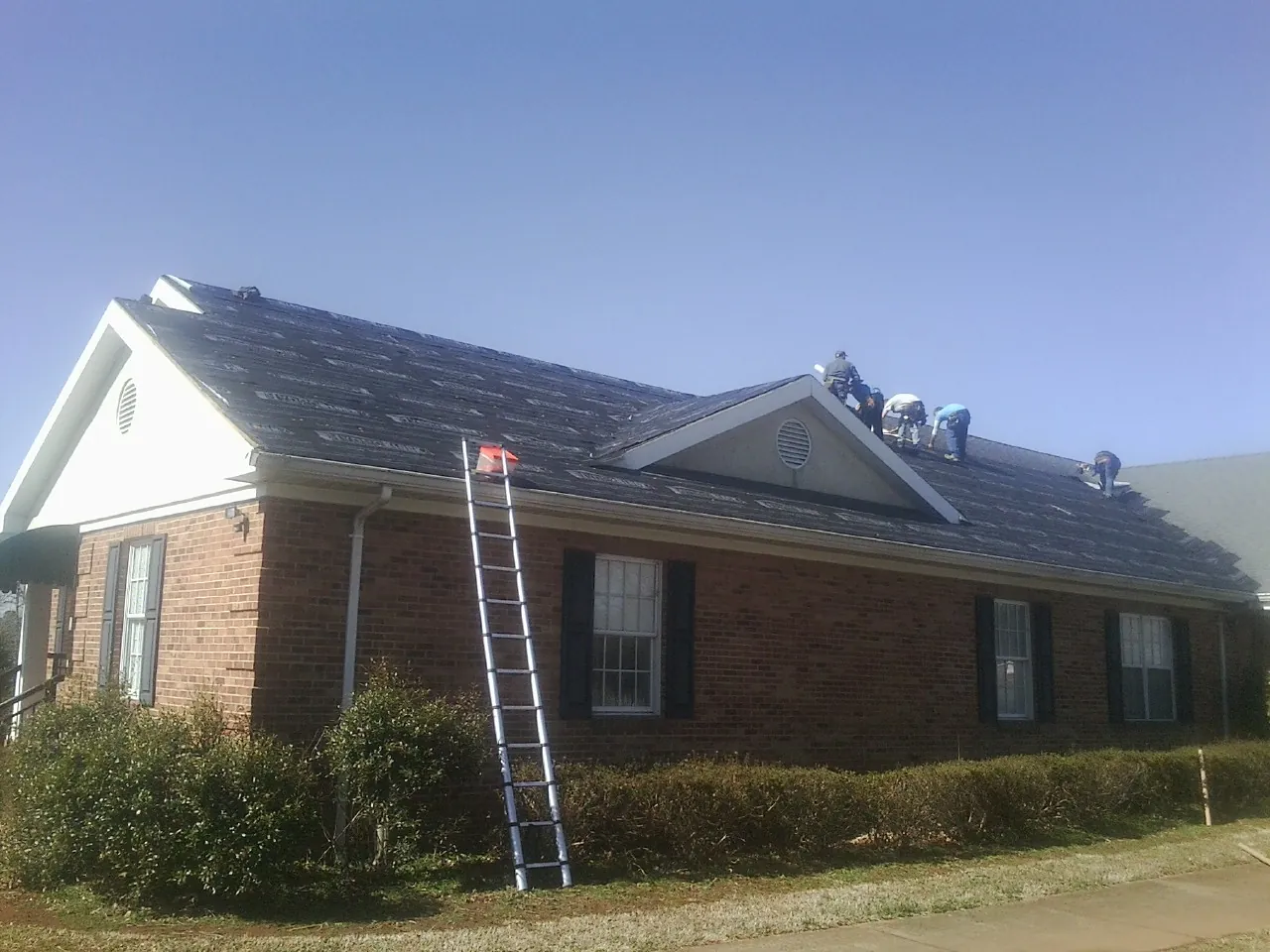 Crew tearing shingles off a brick ranch roof with a ladder set — Emergency Roof Repair work by our crew in Glen Head