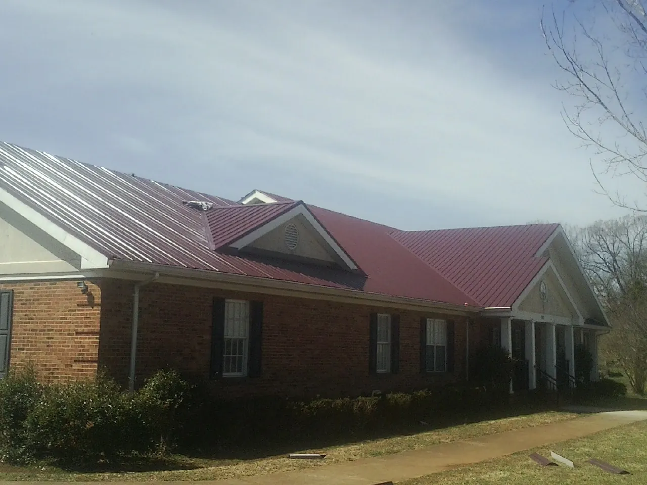 Red metal roof with gabled entries on a long brick building, from a Hail Damage Roof Repair job in Glen Head