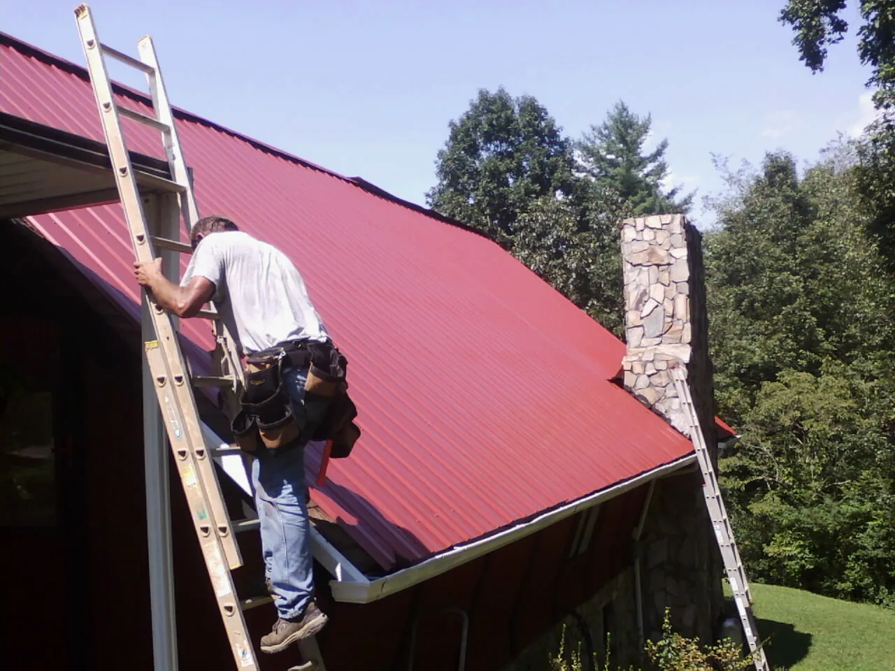 Roofer on a ladder beside a red metal roof and stone chimney — Soffit & Fascia Repair by Roofing Brothers in Glen Head