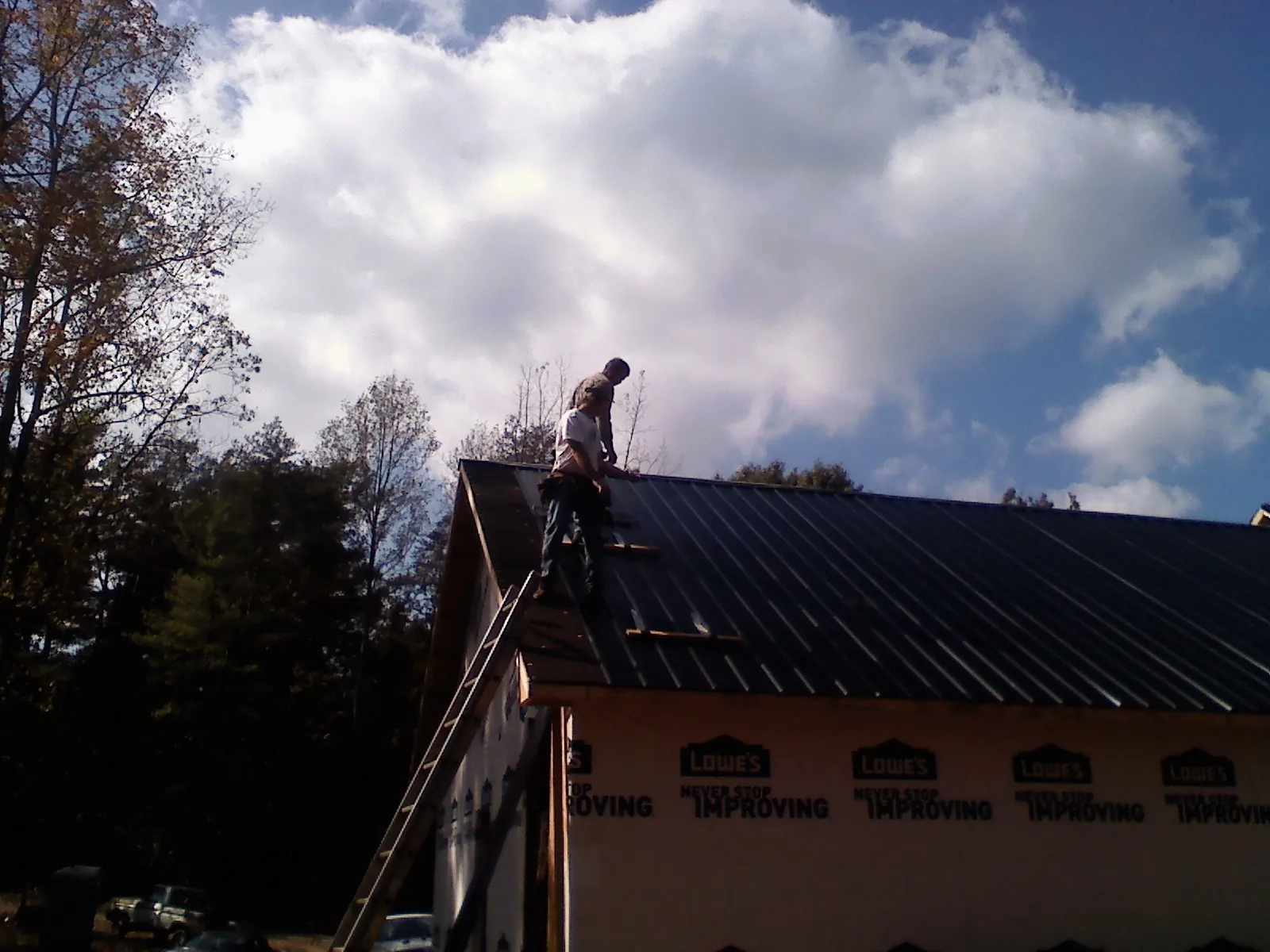 Roofer fastening dark metal panels high against a cloudy sky, from a Roof Inspection job in Glen Head